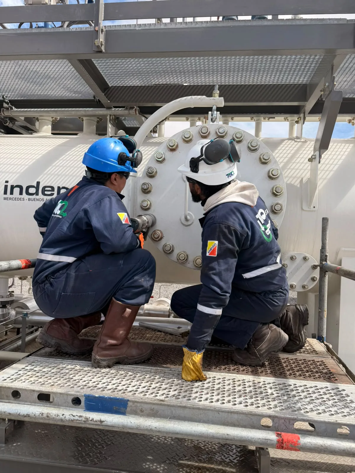 Dos trabajadores con la marca TBQ en su ropa junto a equipamiento industrial.